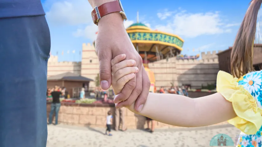 A child is holding hands with an adult and in the background an amusement park
