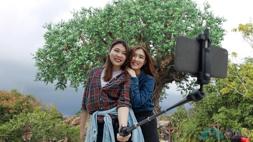 Two friends pose for photo with a selfie stick in front of Disney's Animal Kingdom Park tree in Orlando