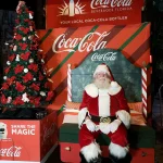 Papai Noel sentado ao lado de uma árvore de Natal com emblema da Coca-Cola ao fundo