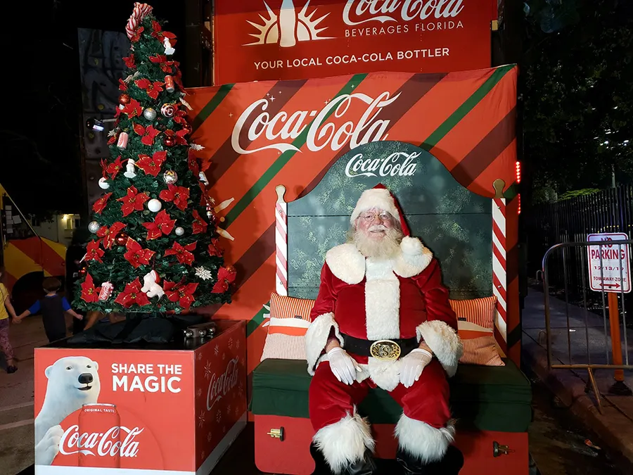 Papai Noel sentado ao lado de uma árvore de Natal com emblema da Coca-Cola ao fundo